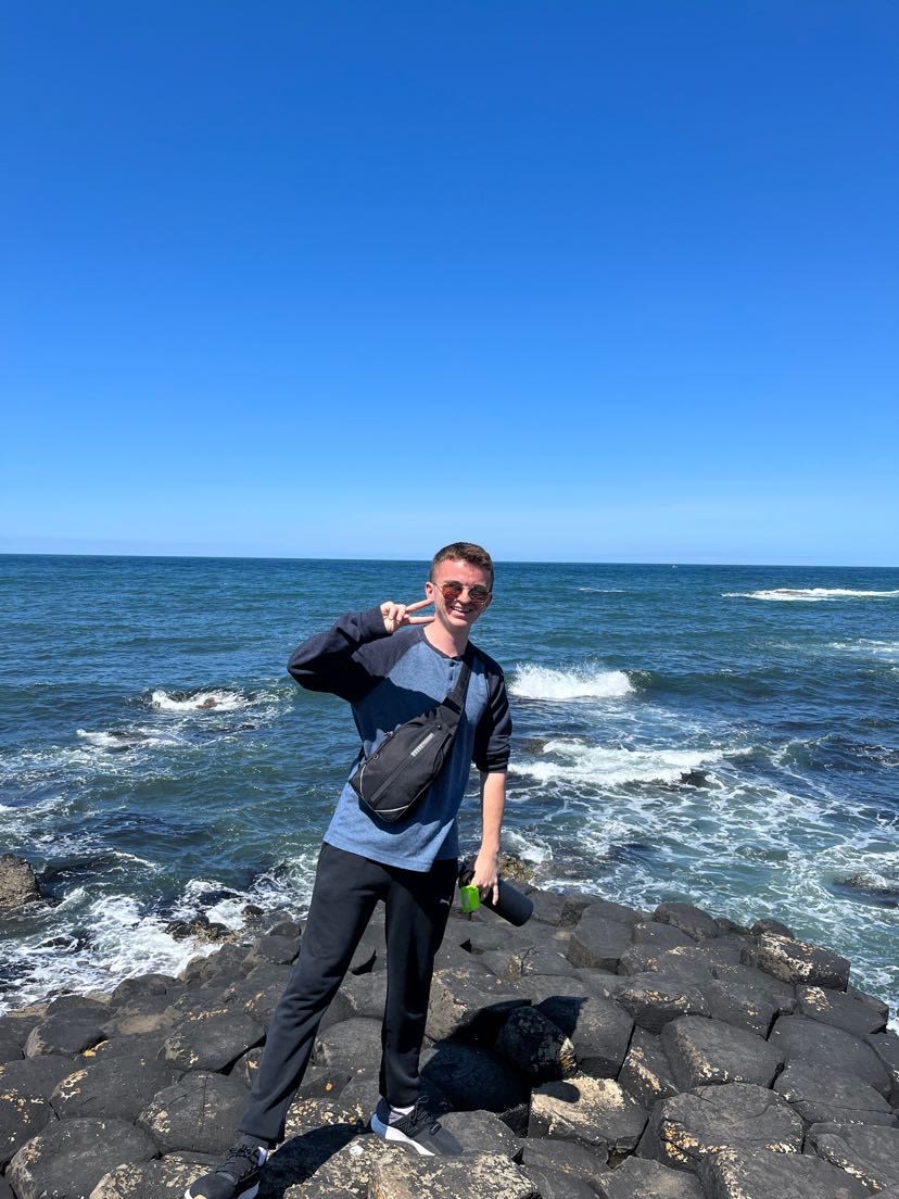 Team member, Andrew "Perky" Poerkson poses on black rocks with a blue ocean and blue skies in the background.