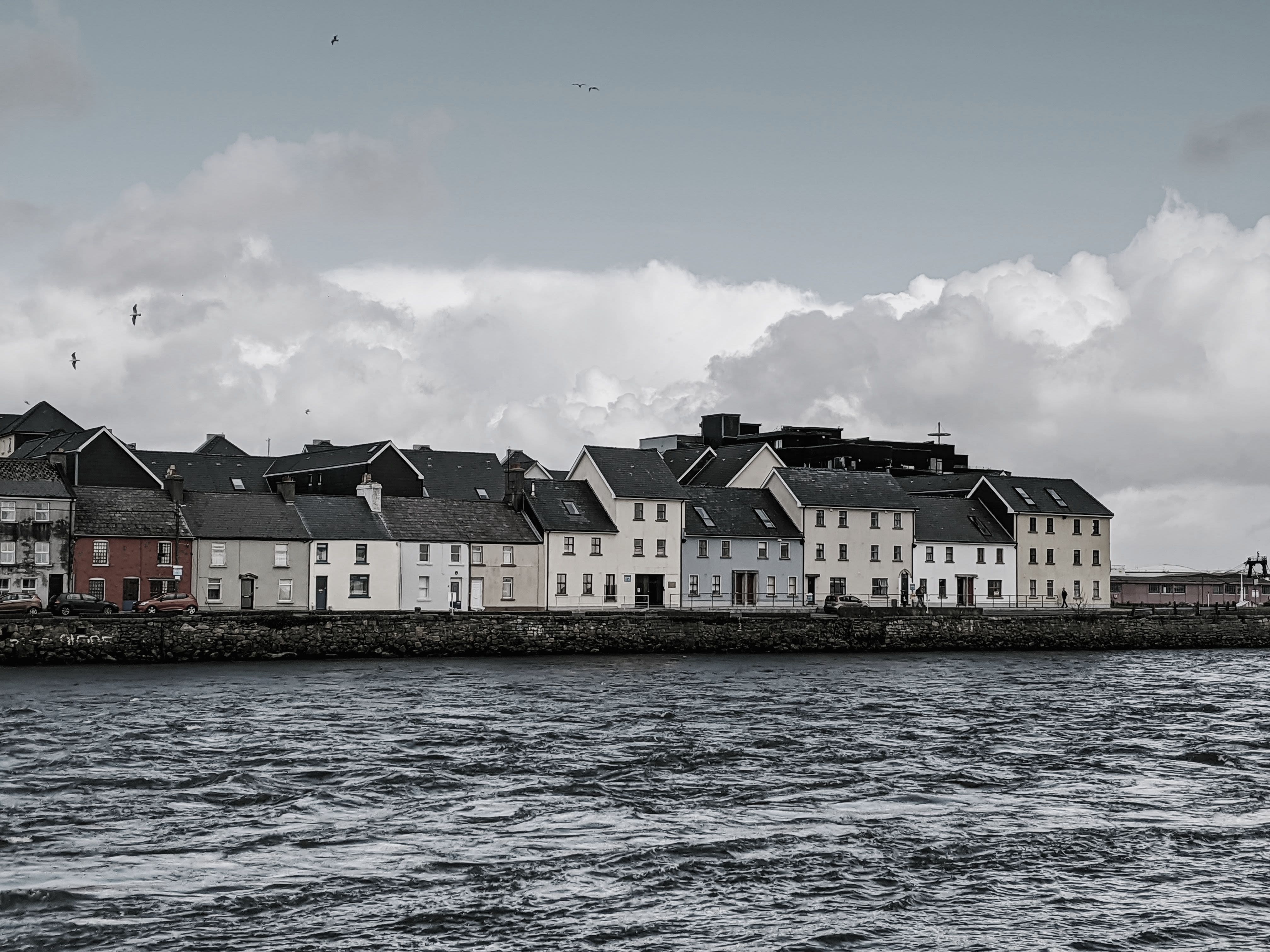 A view across the water. There are multiple connected buildings that are all different colors with clouds in the background.